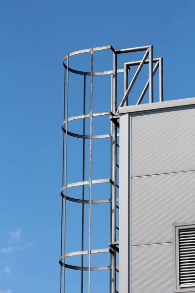 Side view of metal vertical access ladders with round safety tunnel for use as emergency fire escape in case of fire mounted on side of industrial building at local industrial complex on clear blue sky background