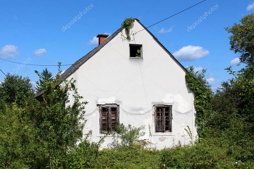 Side view of abandoned small white suburban family house with broken windows and dilapidated facade covered with old roof tiles completely surrounded with overgrown crawler plants and various vegetation on partially cloudy blue sky background