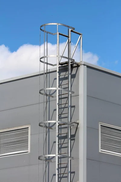 Vertical access ladders with round safety tunnel for use as emergency fire escape in case of fire mounted on side of new industrial building at local industrial complex on partially cloudy blue sky background