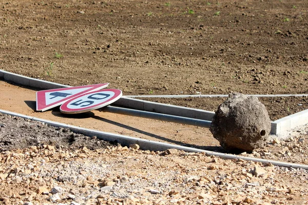 Knocked over old road sign for 50 speed limit and triangle traffic merges ahead onto main carriageway road sign mounted on metal pole with large concrete foundation left at local construction site surrounded with concrete curb and sand