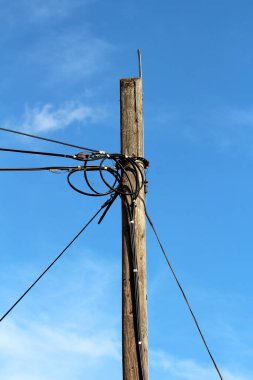 Paslı tutuculara bağlı birden fazla elektrik kablosu olan klasik ahşap elektrik direği kısmen bulutlu mavi gökyüzü arka planında her yöne gidiyor.