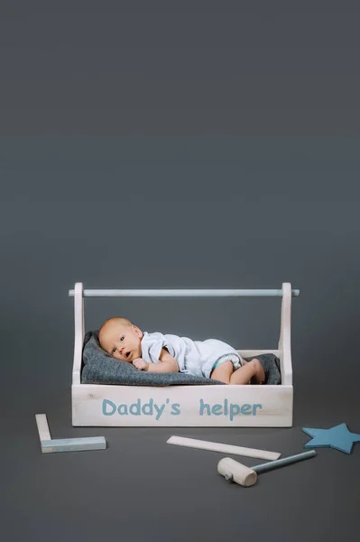 Little baby in bodysuit lying in wooden toolkit with daddys helper lettering and hammer near by — Stock Photo