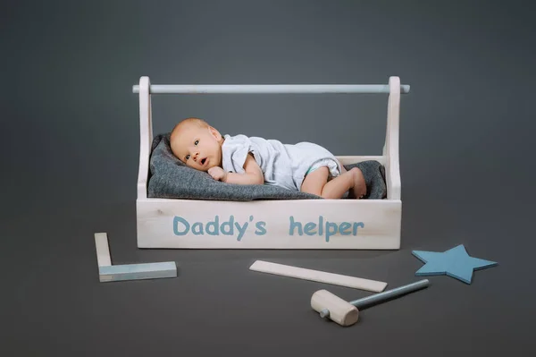 Little baby in bodysuit lying in wooden toolkit with daddys helper lettering and hammer near by — Stock Photo
