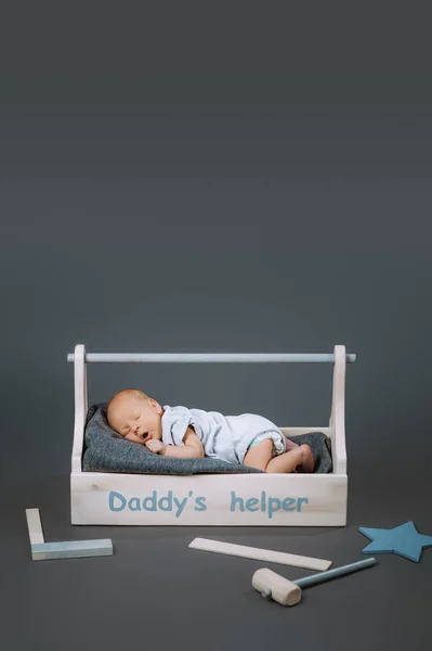 Petit bébé en costume de corps bâillant dans une boîte à outils en bois avec des papas aide lettrage et marteau à proximité — Photo de stock