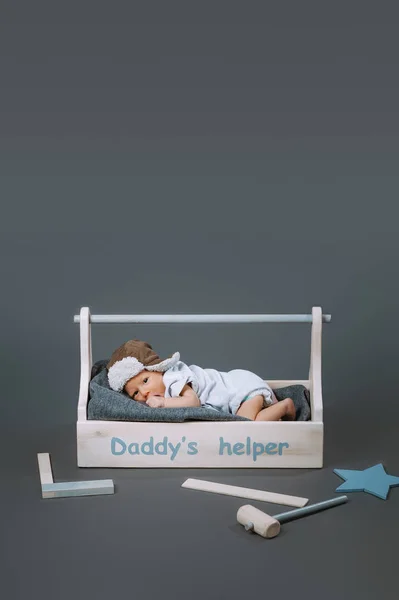 Petit bébé en body couché dans une boîte à outils en bois avec lettrage daddys helper et marteau à proximité — Photo de stock