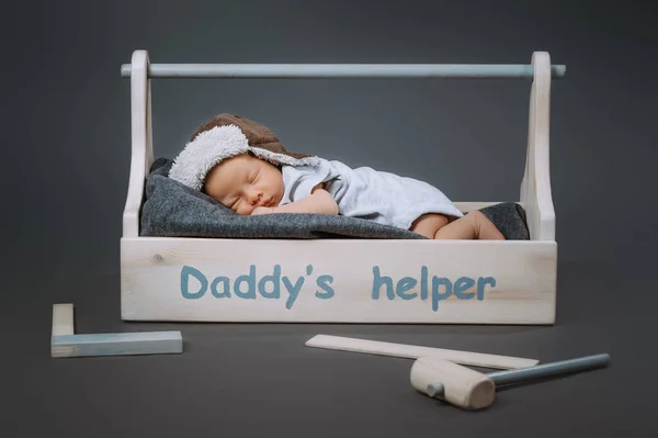 Adorable newborn baby in hat sleeping in wooden toolkit with daddys helper lettering and hammer near by — Stock Photo