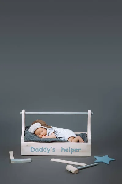 Petit bébé en body couché dans une boîte à outils en bois avec lettrage daddys helper et marteau à proximité — Photo de stock