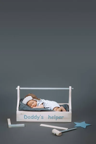 Pequeño bebé en traje de cuerpo acostado en kit de herramientas de madera con daddys letras ayudante y martillo cerca de - foto de stock