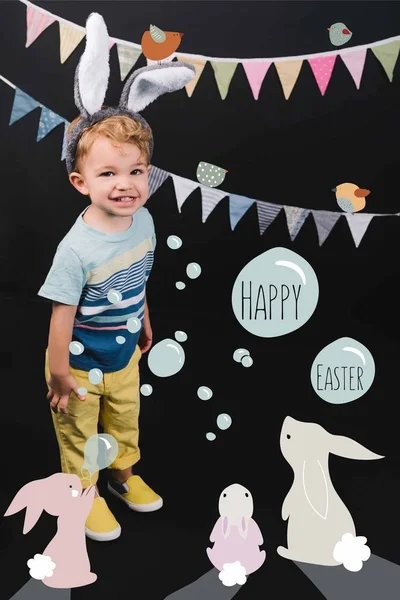 Adorable niño pequeño con orejas de conejito y guirnalda sonriendo a la cámara en negro, feliz Pascua letras en burbujas y conejitos collage - foto de stock