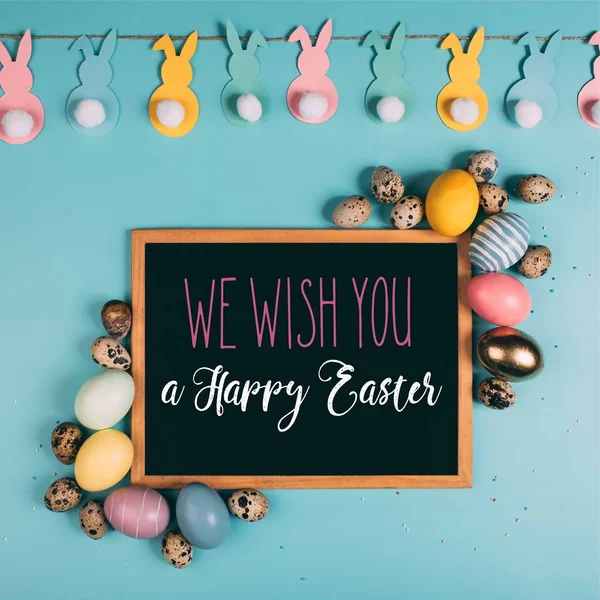 Top view of colorful easter eggs and we wish you happy easter lettering chalkboard on blue surface — Stock Photo