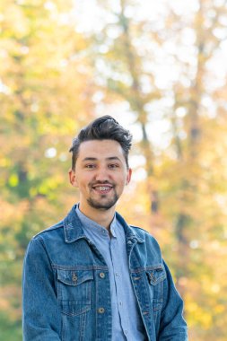 Yakışıklı genç adam kameraya bakıyor. Dışarıda kot ceketli, mavi gömlekli, kendinden emin ve başarılı bir gencin portresi. Mutlu adam gülümsüyor.