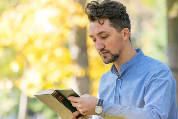 Handsome young man reading a book in a park. Portrait of a young man with a denim jacket and blue shirt reading a book outside. A guy reading a book in a park.