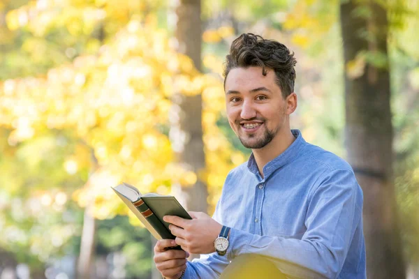Handsome young man reading a book in a park. Portrait of a young man with a denim jacket and blue shirt reading a book outside. A guy reading a book in a park.