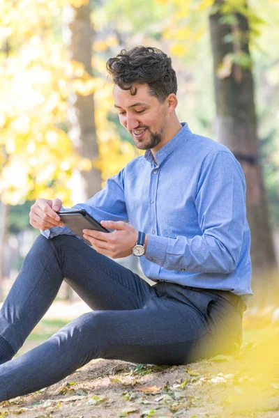 Handsome young man reading an ebook in a park. Portrait of a young man with a denim jacket and blue shirt reading an e-book outside. A guy reading an ebook in a park.