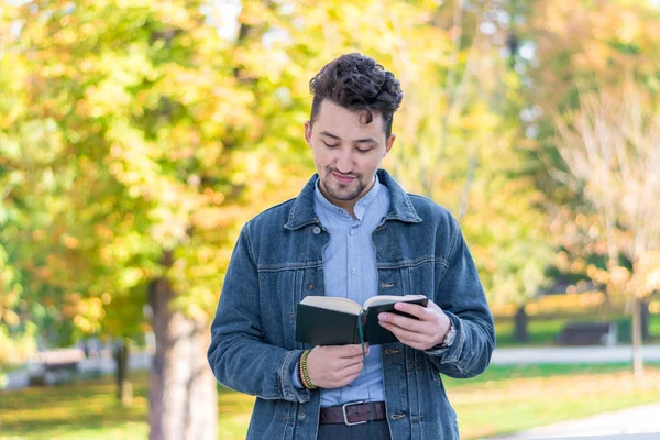 Handsome young man reading a book in a park. Portrait of a young man with a denim jacket and blue shirt reading a book outside. A guy reading a book in a park.