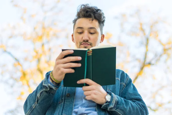 Handsome young man reading a book in a park. Portrait of a young man with a denim jacket and blue shirt reading a book outside. A guy reading a book in a park.