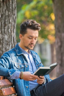 Parkta kitap okuyan yakışıklı bir delikanlı. Dışarıda kitap okuyan kot ceketli ve mavi gömlekli genç bir adamın portresi. Parkta kitap okuyan bir adam..
