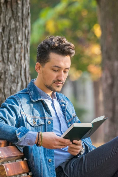 Handsome young man reading a book in a park. Portrait of a young man with a denim jacket and blue shirt reading a book outside. A guy reading a book in a park.