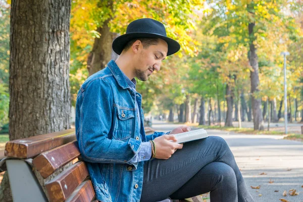 Handsome young man reading a book in a park. Portrait of a young man with a denim jacket and blue shirt reading a book outside. A guy reading a book in a park.