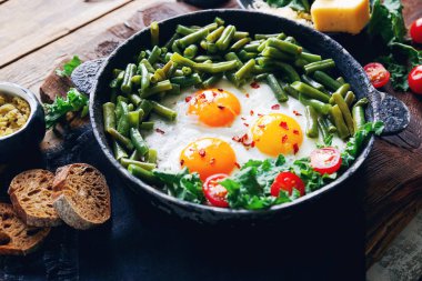 Domatesli bir kap taze yumurta, yeşil fasulye, ekmek, baharat ve otlar kırsal ahşap arka planda, sağlıklı kahvaltı kavramı, seçici odaklanma.