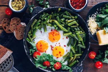 Domatesli bir kap taze yumurta, yeşil fasulye, ekmek, baharat ve otlar kırsal ahşap arka planda, sağlıklı kahvaltı konsepti, üst manzara.