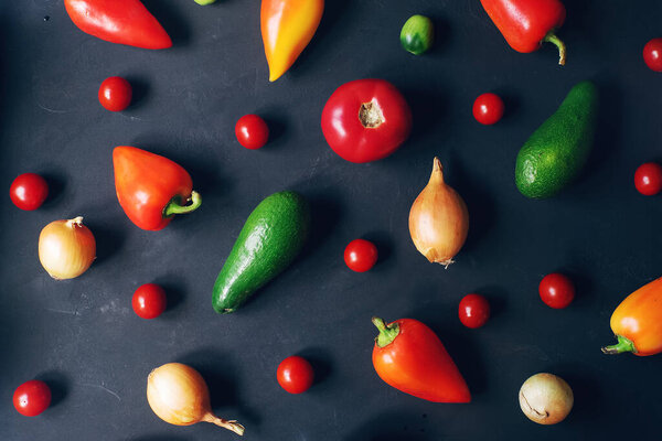 Pattern of raw fresh cherry tomatoes, pepper, avocado and onion on dark stone table, organic food concept, top view