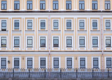 Önünde klasik metal çit olan sarı cepheli klasik bina..