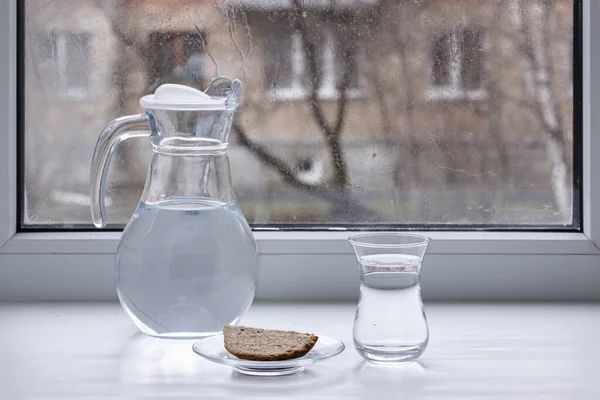 A carafe of water, a glass and a slice of bread in front of the window.