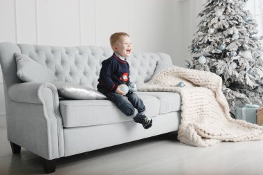 Küçük çocuk noel ağacının yanındaki kanepede oturuyor ve çok gülümsüyor.