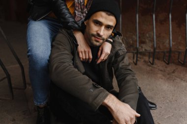 Sakallı yakışıklı bir adam yolda oturuyor ve kadın elleri omuzlarında. Erkek portresi