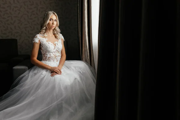 Elegantní blondýny nevěsta ve svatebních šatech sedí před okny — Stock fotografie