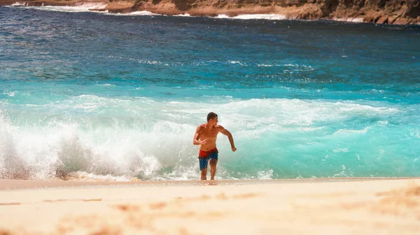 The guy is running away from the waves of the ocean in shorts on the beach sun heat — Zdjęcie stockowe