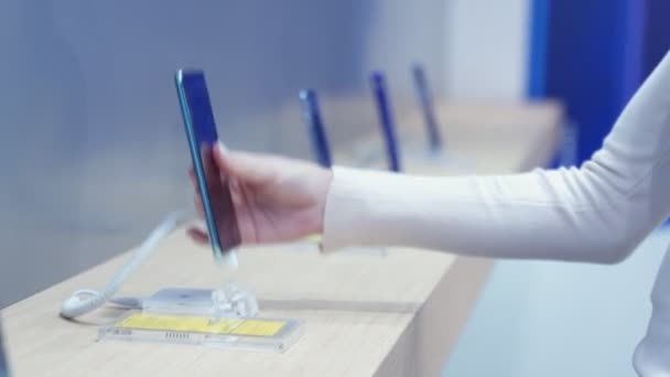 Mujer elige un teléfono inteligente en una tienda de electrónica, de cerca. Ella toma el teléfono inteligente del mostrador e intenta usarlo . — Vídeo de stock