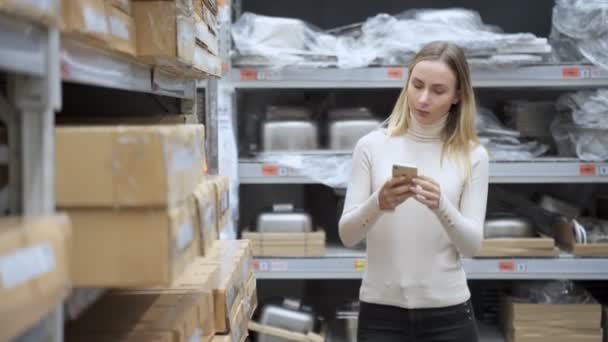 Женщина с телефоном в магазине на складе. Молодая женщина ищет товары на полках с помощью смартфона . — стоковое видео