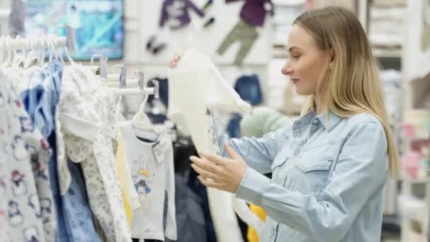 Hermosa chica eligiendo ropa para el bebé de pie cerca de un estante de ropa para niños en el supermercado, compras, almacén del centro comercial . — Vídeos de Stock