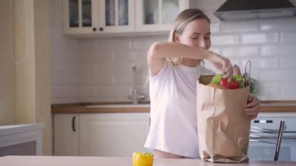 Feliz de relajar a la joven mujer sosteniendo la bolsa de papel de comestibles en la cocina. Una mujer desempaca verduras y frutas de una bolsa de supermercado . — Vídeos de Stock