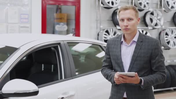 Muž prodávající s tabletem ukazuje auto v showroomu. Úspěšný prodejce stojící před moderním vozidlem předvádí ukázky aut a pohledů do kamery s úsměvem. Koncept prodeje automobilů — Stock video