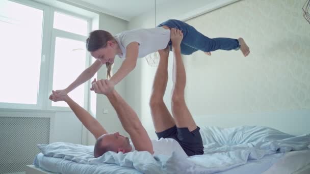 Glada par som leker i sängen och har roligt. Mannen som ligger på sängen och lyfter sin fru med fötterna i handen. — Stockvideo