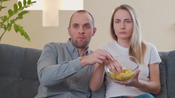 한 부부가 간식을 먹으면서 소파에서 무서운 영화를 보고 있다 — 비디오