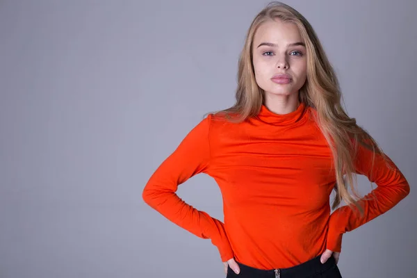 Schönes Porträt eines gut gekleideten modischen Mädchens in orangefarbenem Sweatshirt. Schöne Dame posiert drinnen auf grauem Hintergrund. Blondes Model mit ernstem Blick. Mode- und Lifestyle-Konzept — Stockfoto