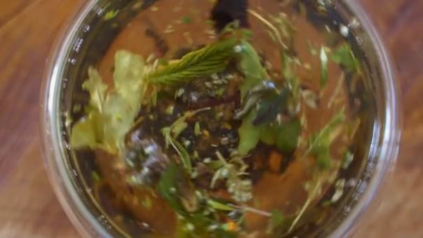 Vista dall'alto primo piano foglie di tè che si aprono in acqua calda a tazza trasparente. Preparazione di tisane verdi o nere in tazza ad acqua bollita — Video Stock