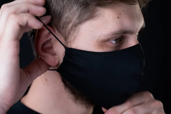 Der Mann setzt sich eine Gesichtsmaske auf. Schwarze, stylische Gesichtsmaske während einer Virenpandemie. Handgemachte Gesichtsmaske aus Baumwolle zum Schutz vor Viren und Bakterien. — Stockfoto