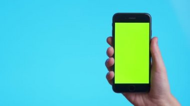 Woman hand holds a smartphone with greeen screen over a blue background. Close up footage of hand only