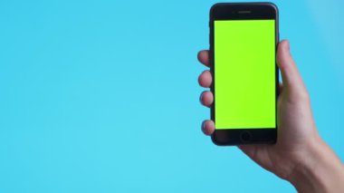 Woman hand holds a smartphone with greeen screen over a blue background. Close up footage of hand only