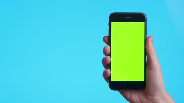 Woman hand holds a smartphone with greeen screen over a blue background. Close up footage of hand only — Stok video
