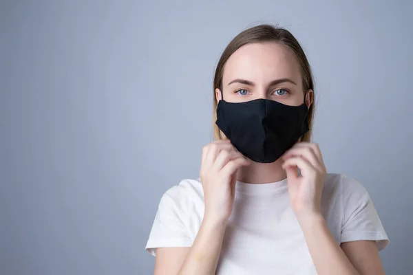 Mulher coloca uma máscara médica preta em seu rosto em um fundo cinza — Fotografia de Stock