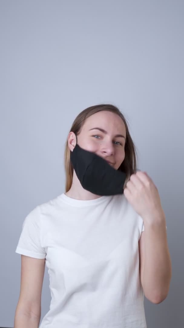 Vídeo vertical. El retrato de cerca de una mujer se quita una máscara facial médica negra durante la cuarentena del coronavirus COVID-19. Sobre un fondo gris — Vídeo de stock