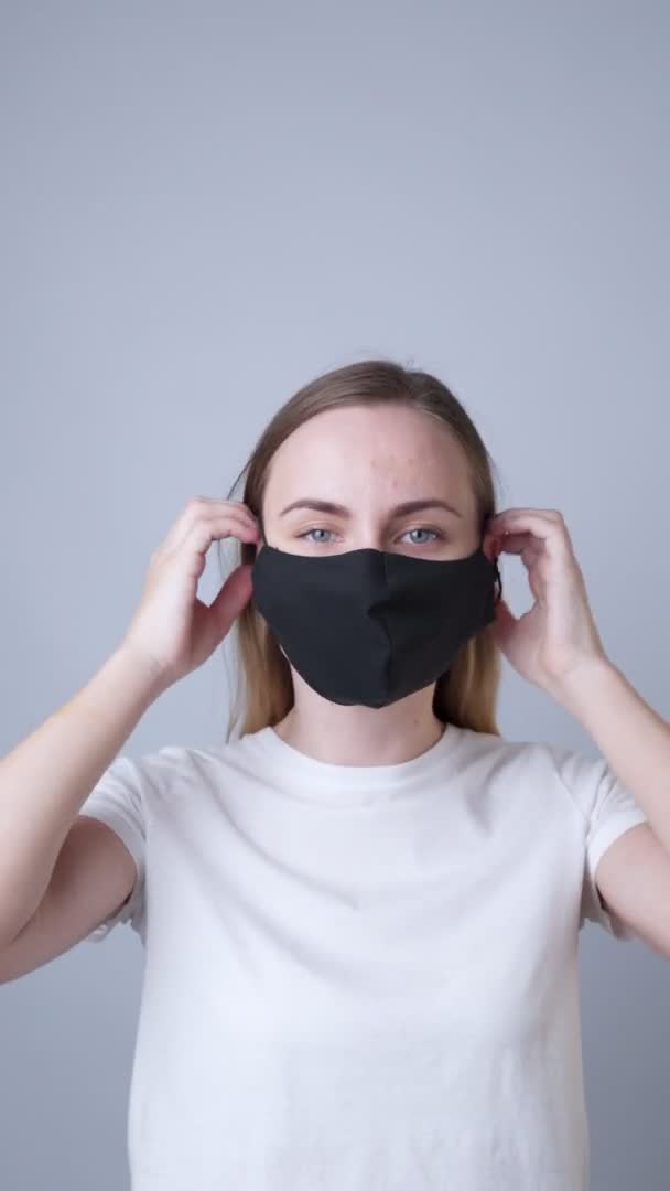 Вертикальное видео. Женщина надевает черную медицинскую маску на лицо на сером фоне — стоковое видео