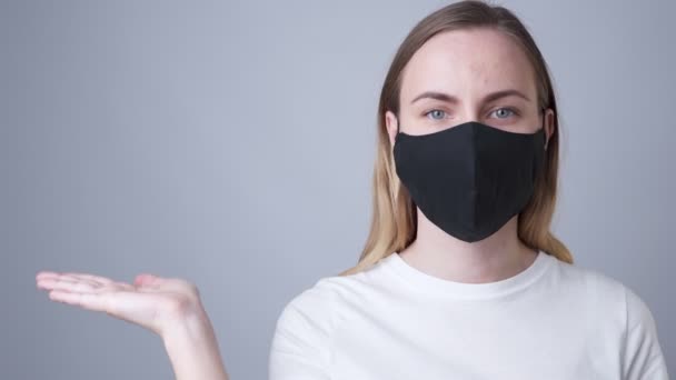 Mujer en una máscara médica negra mostrando y presentando con la palma de la mano abierta sobre un fondo gris . — Vídeo de stock