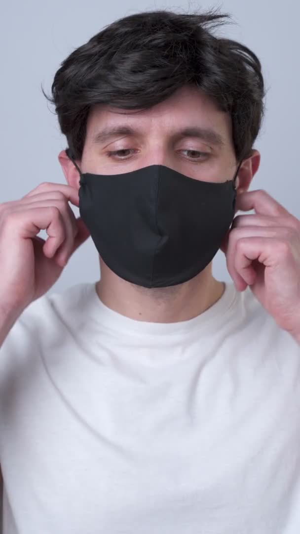 Вертикальне відео. Збільшений портрет людини знімає чорну медичну маску під час карантину вірусу COVID-19. На сірому фоні — стокове відео